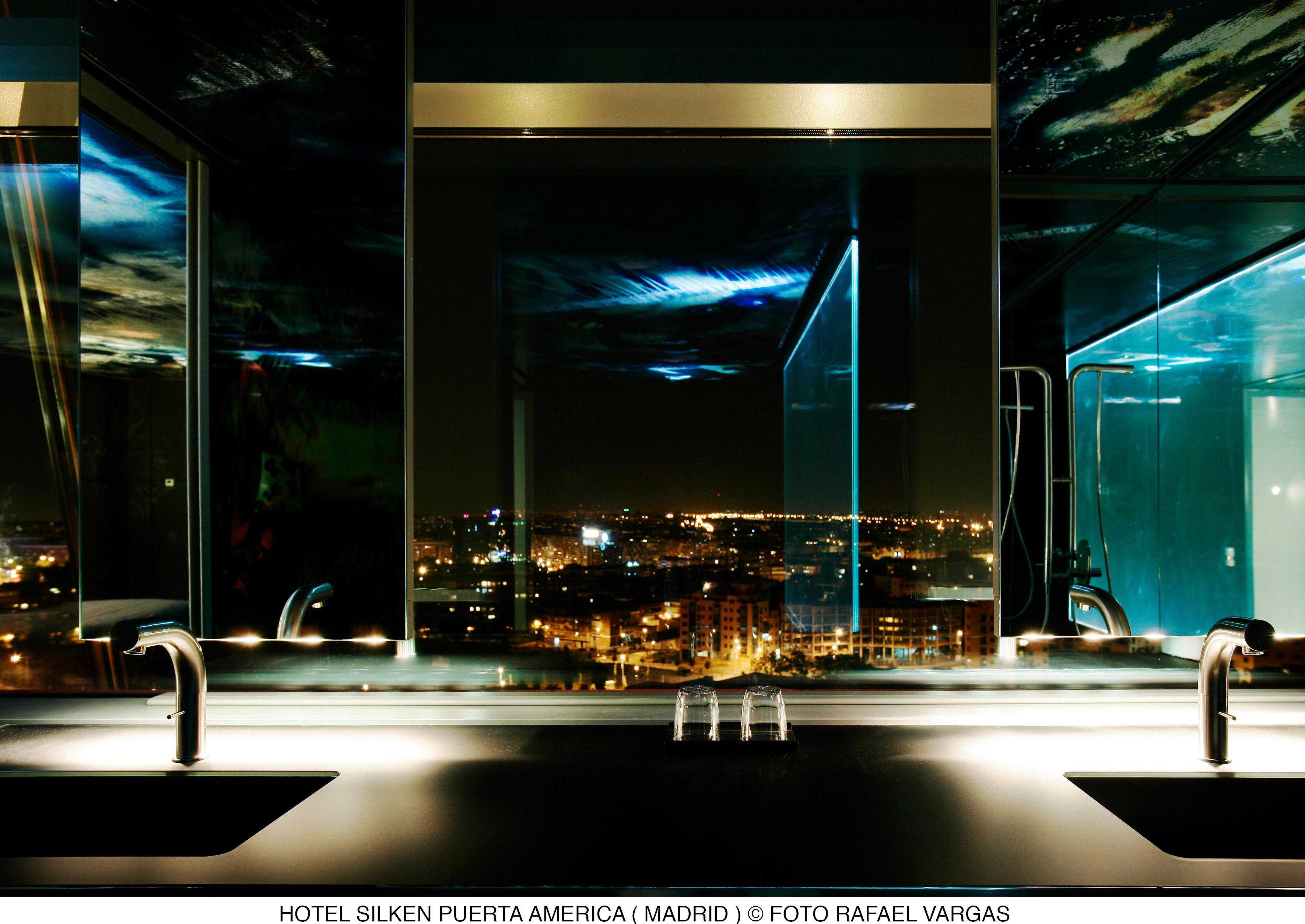 Hotel Puerta America Madrid Esterno foto The photo showcases a modern bathroom with a sleek design, featuring two sinks with contemporary faucets. The large mirrors reflect the city skyline at night, illuminated by various lights from buildings and streets below. The bathroom has a clean, m